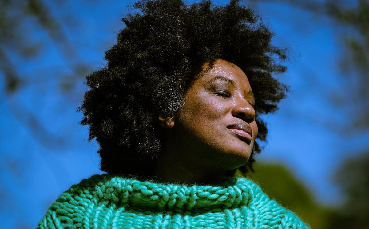 Headshot of Aja Barber wearing a teal knit jumper with branches and the sky in the background.