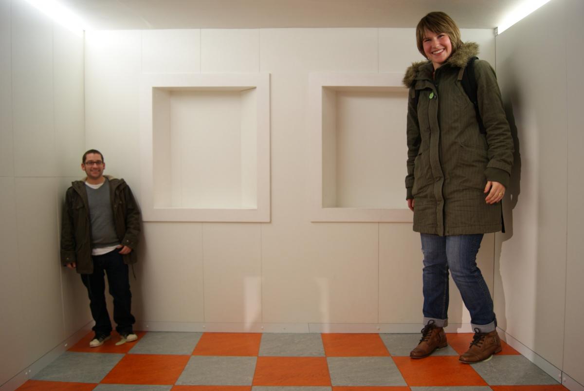 A woman standing to the far right seeming very tall almost touching the ceiling and a man standing to the far left seeming very short demonstrating the Ames room illusion. 
