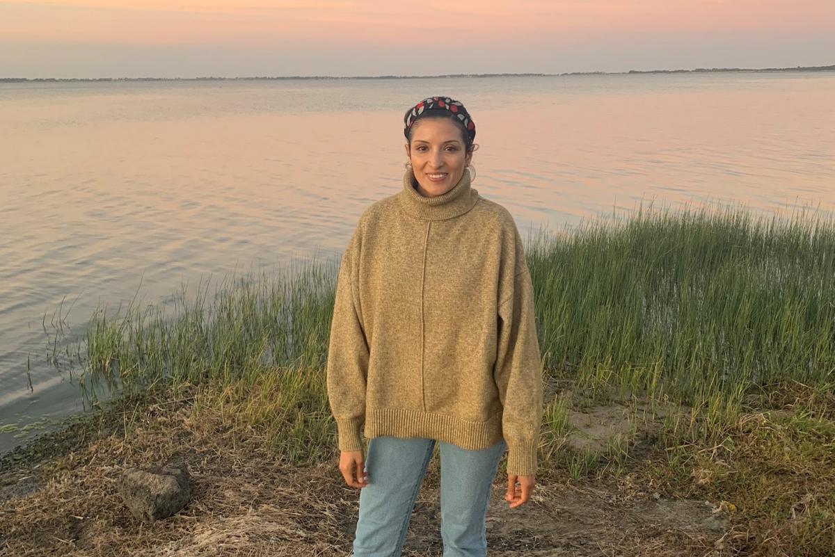 Carolina Mayen in a beige sweater and blue jeans near a lake, smiling at the camera.