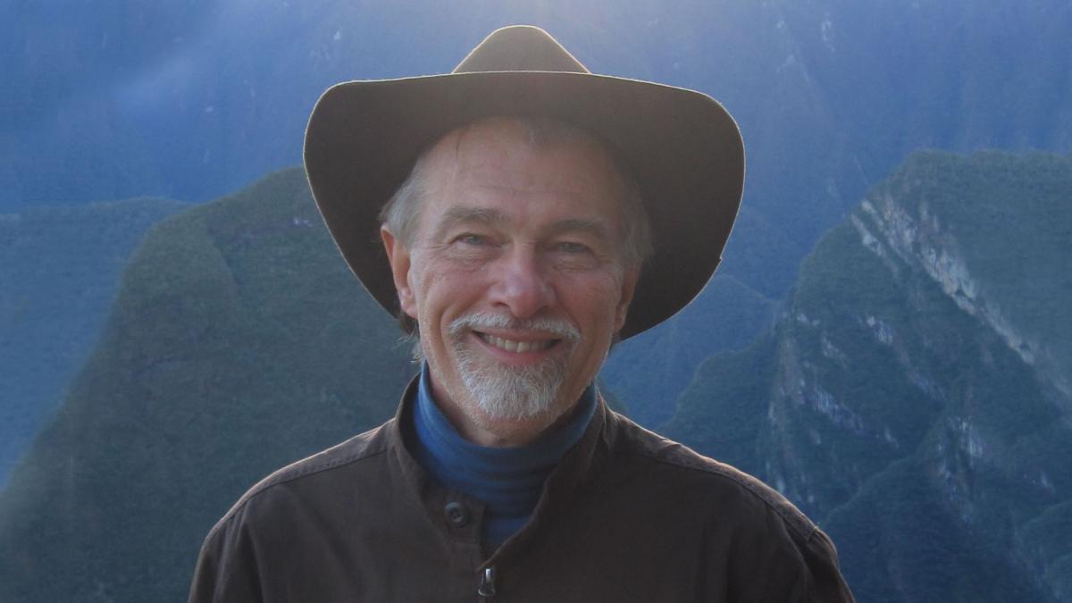 Michael Winkelman is standing in front of a scenic mountain range. He is wearing a brown hat, a blur turtle neck and over it a brown sweater. 