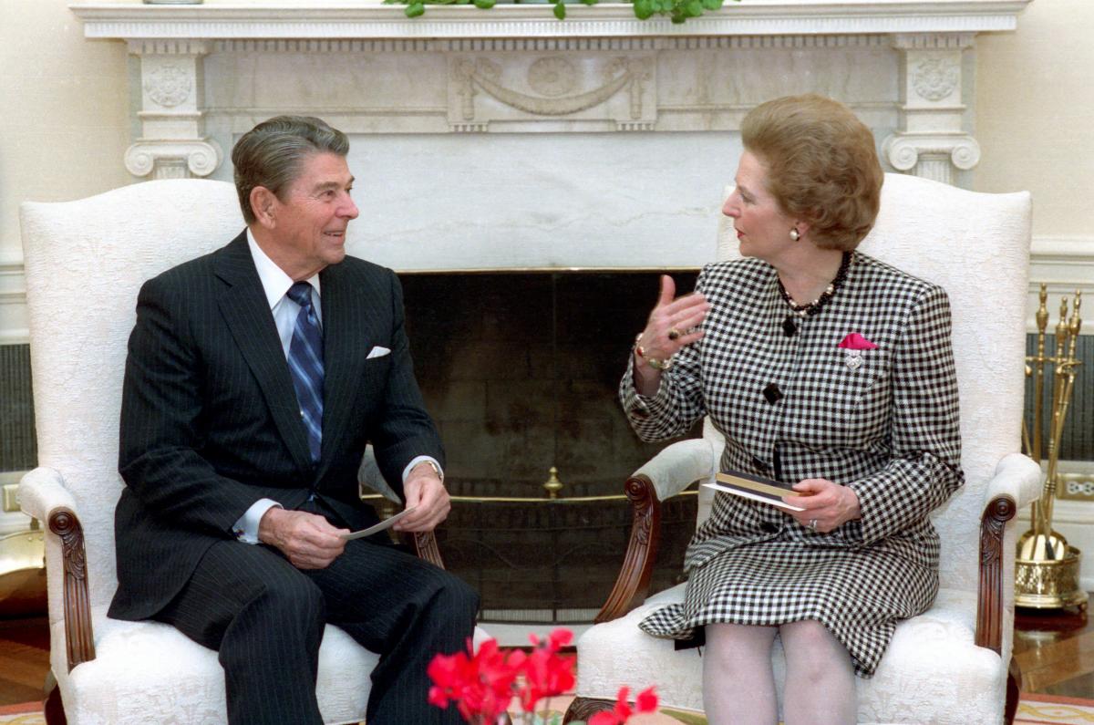 Picture of Ronald Reagan & Margaret Thatcher, sitting in next to each other talking. Margaret wears a grey checkered suit and skirt and Ronald wears a black striped  suit