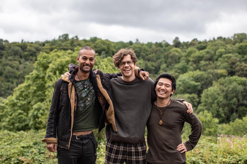 Simon Ruffell, with Nige Netzband on the left, and WaiFung Tsang on the right, standing with their arms on each others shoulders with a forested area behind them and smiling at the camera.