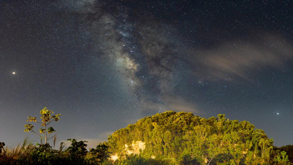 A view of the night sky with the moon, stars and muted colours over the lush green mountain