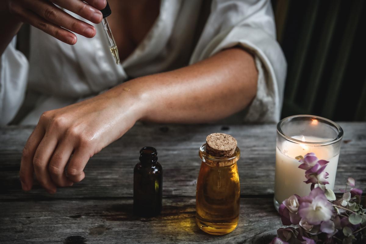 A person using a dropper to drip essential oil to massage themselves. In front of them on a table, a candle, a body oil in a glass jar and an open essential oil bottle is placed. 