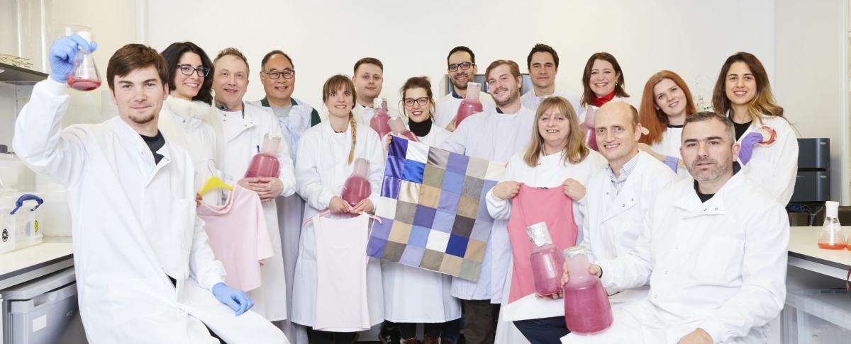 The Colorifix team wearing lab coats holding conical flasks with pink dye blend along with a blue patchwork fabric.