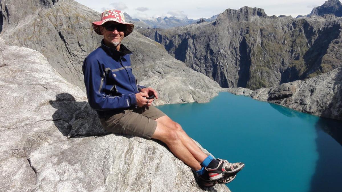 Photograph of Dave Vass wearing a blue jacket, brown shorts and a hat, while sitting on a rocky mountain.