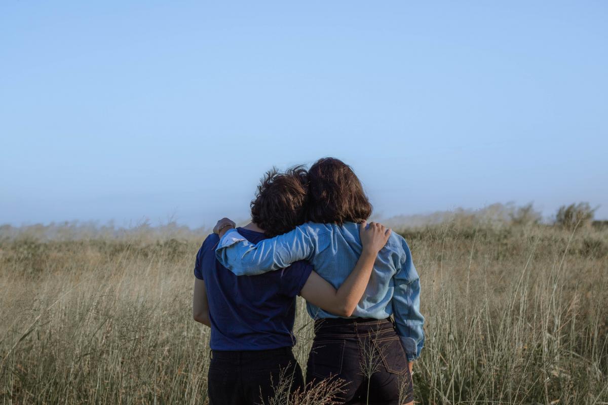 Two people with their backs to the camera leaning on each other with their arms on each others' shoulders standing in a field.