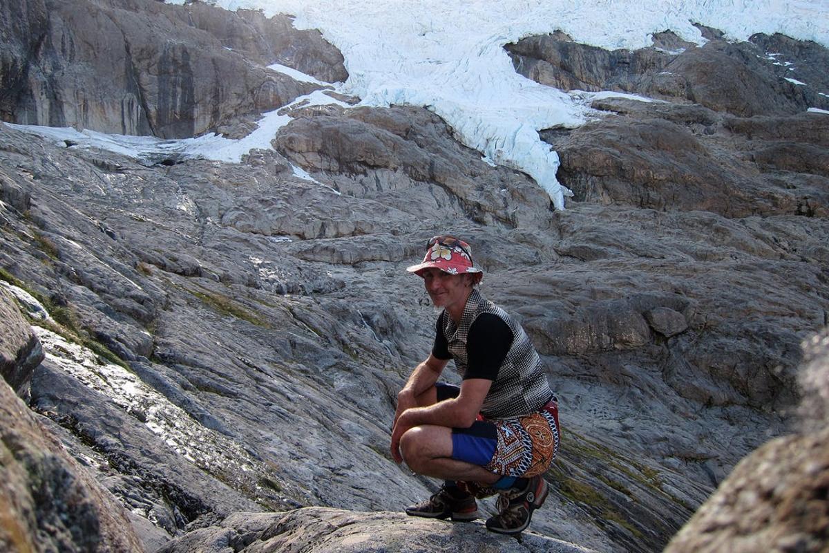 Dave Vass sitting on a rocky mountain, smiling at the camera, wearing a floral hat, checkered shirt and shorts