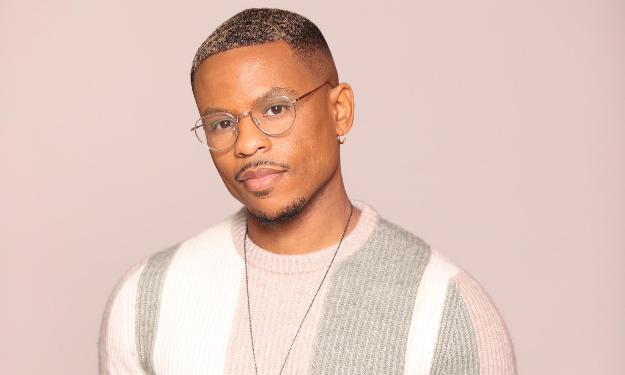 Yolo Akili Robinson wearing a sweater of pink, white, and grey stripes and metal rimmed glasses, standing and looking at the camera.