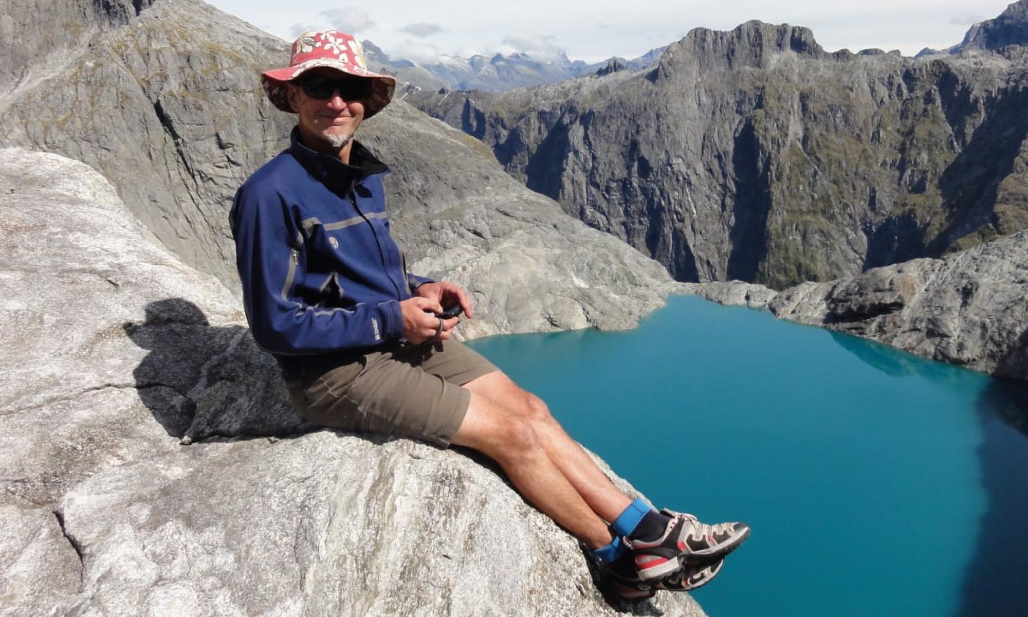 Photograph of Dave Vass wearing a blue jacket, brown shorts and a hat, while sitting on a rocky mountain.